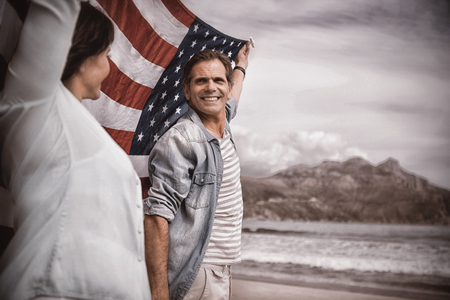 Mature couple holding American flag on beach against skyの写真素材