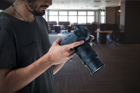 Young photographer operating digital camera  against table and empty chairs in office