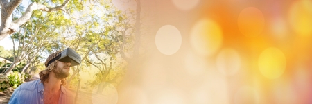 Digital composite of Man in forest with virtual reality headset looking up with orange bokeh...