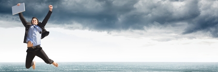 Digital composite of Business man with laptop celebrating against grey clouds and waterの写真素材
