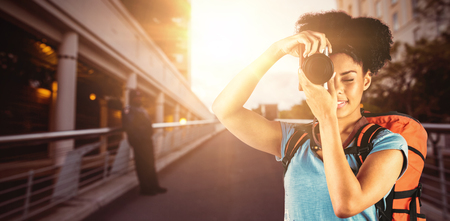 Young woman with backpack taking picture against footbridge against sky in cityの写真素材
