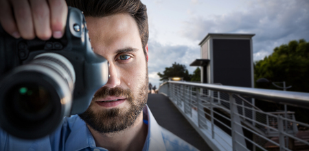 3D Portrait of male photographer photographing through camera against footbridge against cloudy skyの写真素材