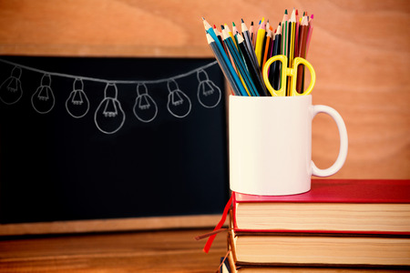 Illustration image of light bulbs arranged against desk organizer on stack of books against blackboardの写真素材