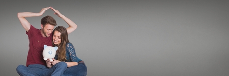 Digital composite of Happy couple sitting and holding a piggy bank against grey backgroundの写真素材