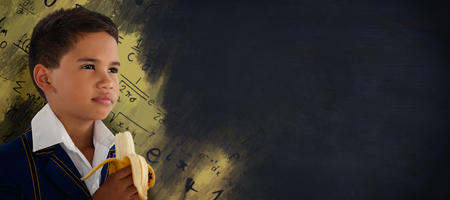 Schoolboy looking away while eating banana against black backgroundの写真素材