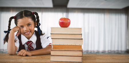 Schoolgirl leaning by books and apple on desk against spacious room with curtains on windowの写真素材