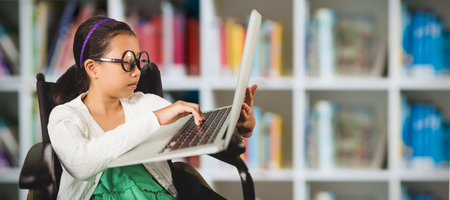 Young girl typing on laptop while sitting against defocused books on shelfの写真素材