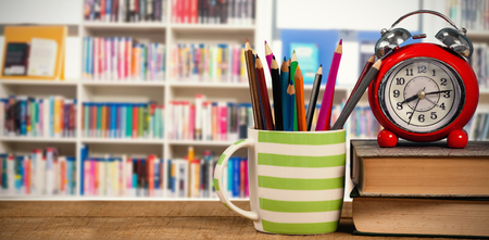 Books with alarm clock by colored pencils in mug on table against various books on shelf in libraryの写真素材