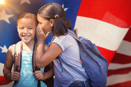 Girl with backpack whispering in friend ear against american flag with stripes and starsの写真素材