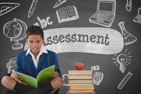Digital composite of Happy student boy at table reading against grey blackboard with assessment text and education and scの写真素材