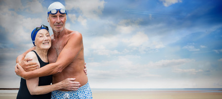 Smiling senior couple in swimwear embracing while standing against serene beach landscapeの写真素材