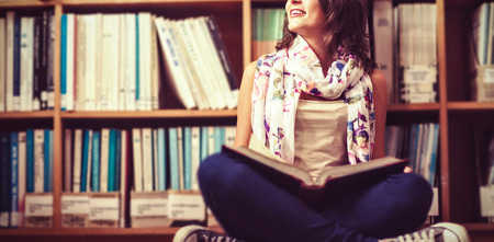 Thoughtful female student sitting against bookshelf with book on library floor at college