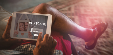 Graphic image of mortgage login page and couple holding model house against low section of man using digital tablet while sitting on sofa in living roomの写真素材