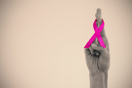Cropped hand showing crossed fingers with pink ribbon against gray backgroundの写真素材