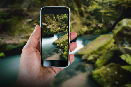 Cropped hand holding smart phone against rapids flowing along lush forestの写真素材