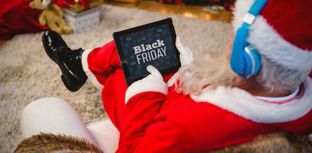 Black Friday text with Christmas icons on blackboard against santa claus using digital tablet at homeの写真素材