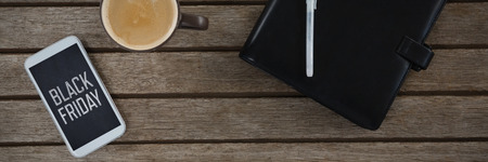 Mobile phone, coffee, pen and organizer on wooden plank against black friday advert
