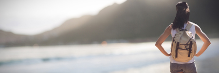 Rear view of female hiker carrying backpack standing with hands on hip against mountain against sky at beachの写真素材