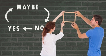 Digital composite of Couple holding picture frame against wall with Yes No Maybe text and arrowsの写真素材