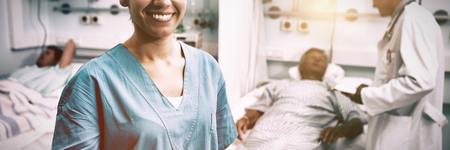 Portrait of happy nurse standing in hospital room with doctor and patient talking in backgroundの写真素材