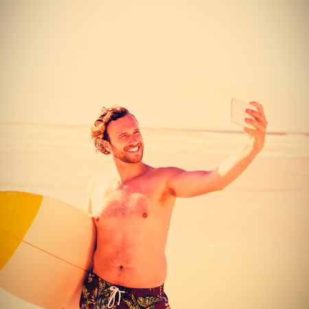 Smiling shirtless man taking selfie with surfboard at beach during sunny day