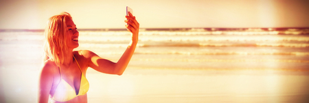 Happy young woman in yellow bikini taking selfie at beach during sunny dayの写真素材