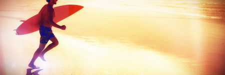 Side view of shirtless man running while holding surfboard at beach during sunny dayの写真素材