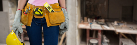 Woman with tool belt and holding hard hat against grey background against workshopの写真素材
