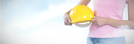 Woman holding hard hat against grey background against blue sky with cloudsの写真素材