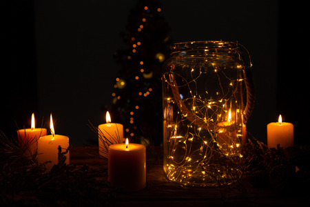 small electric garland in a glass jar with candles and a christmas tree in the backgroundの写真素材