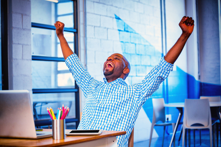 Excited man sitting on table with digital tablet and laptop in officeの写真素材