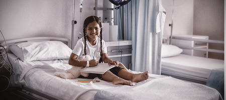 Smiling girl drawing picture in a book while sitting on hospital bedの写真素材