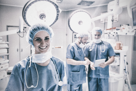 Portrait of smiling female surgeon standing in operation room while colleague discussing background in hospitalの写真素材
