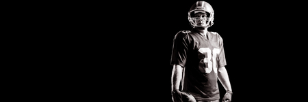 American football player standing with rugby ball and helmat against black backgroundの写真素材