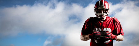 American football player in helmet standing with rugby ball against blue sky with cloudsの写真素材