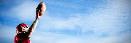 American football player in helmet holding rugby ball with hand raised against low angle view of cloudy skyの写真素材
