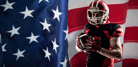 American football player in helmet holding rugby ball against close-up of an american flagの写真素材