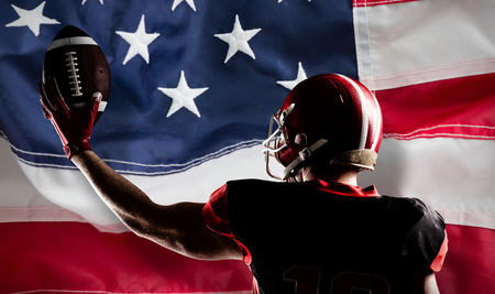 American football player in helmet holding rugby ball against full frame of american flagの写真素材