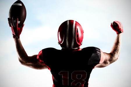 American football player standing with helmet holding football in victory against blue sky with...