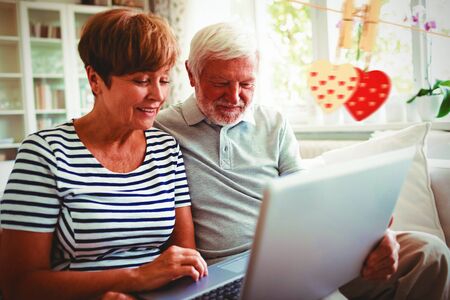 Hearts hanging on a line against senior couple using laptop in living roomの写真素材
