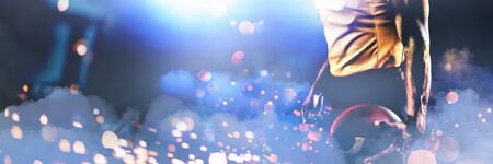 Portrait of confident American football player in helmet holding ball against firework bursting...