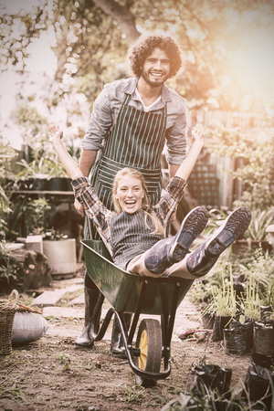 Portrait of cheerful man giving wheelbarrow ride to female gardener outside greenhouseの写真素材