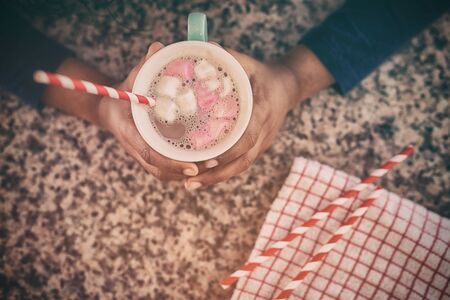 Hands of a young mixed race girl in a kitchen at Christmas, holding a mug with hot chocolate and...