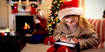 Front view close up of a young Caucasian girl wearing a Santa hat sitting on the sofa using a tablet in her sitting room at Christmas time, with her mother sitting by the fireplace in the backgroundの写真素材