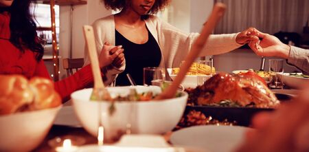Front view of a young adult mixed race woman and a young adult Caucasian woman sitting around a table holding hands with friends saying grace before eating Thanksgiving dinner at home together with friendsの写真素材