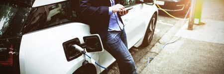 Businessman using mobile phone while charging electric car at charging station