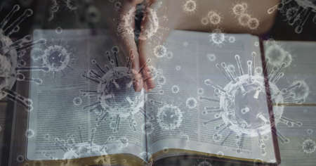 Digital illustration of macro Covid-19 cells floating over Caucasian hands praying over Bible. Religious faith in time of coronavirus global pandemic concept, digital composite.の写真素材