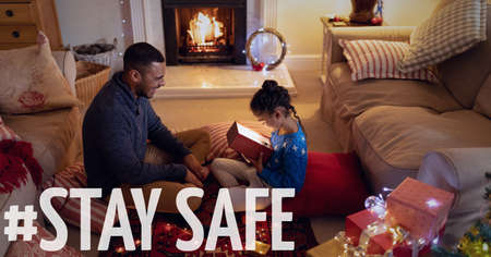 Stay safe text over father with daughter opening present at christmas. covid 19 pandemic and social distancing concept digitally generated image.の写真素材