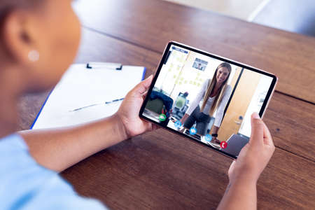 Mixed race businesswoman sitting at desk using tablet having video call with female colleague. work...
