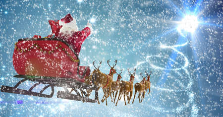 Composition of santa claus in sleigh pulled by reindeer with snow falling on blue background. christmas tradition concept digitally generated image.の写真素材